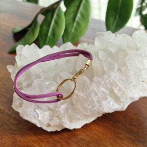 Bracelet double en faux daim mauve, orné d’une breloque ovale et d’un mousqueton en plaqué flash or 18K. Présenté sur fond neutre. Disponible en 15 coloris pour convenir à toutes les envies.