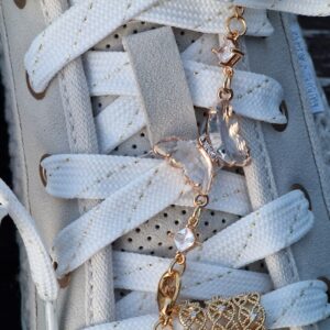 Bijou doré composé d’un papillon en strass blanc encadré par deux strass carrés, suspendu à une chaînette avec mousqueton. Présenté sur une basket Mustang avec éléments décoratifs.